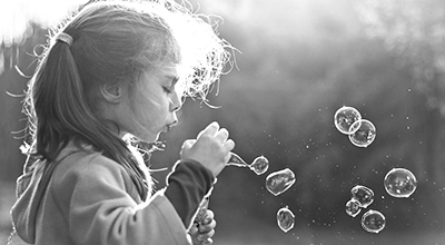 Young girl blowing into a ring to make soap bubbles, with a forest in background (black and white)