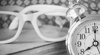Réveille-matin à cloches en avant-plan avec des lunettes à monture blanche posées sur une table de bois (noir et blanc)