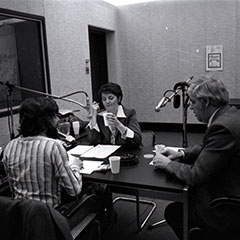 Two men and a woman can be seen behind the microphones of a radio studio.