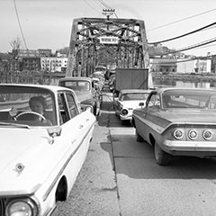 Traffic jam on an iron bridge where a dozen cars are stalled. In the background, we can see a city.