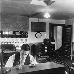 Two men are at the controls of the Shawinigan Water and Power Plant. We can see a communication system in front of them and different control panels in the background.