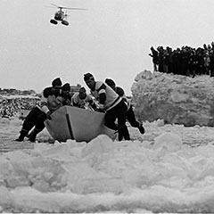 Des hommes tentent de faire avancer un canot sur la glace. À l'arrière d'eux, en hauteur, une foule les encourage.