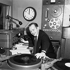 Radio host behind the microphone. There are two turntables, a control board and other studio equipment.