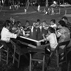 À l'avant-plan, nous pouvons voir deux couples et des enfants mangeant un pique-nique sur une table. À l'arrière, des enfants jouent dans les installations d'un parc pour enfants.