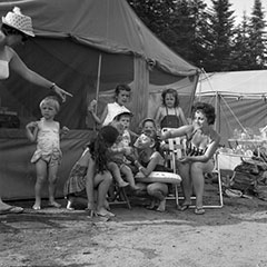 Two mothers and some children are sitting in front of camping tents. They are dressed in their bathing suits.