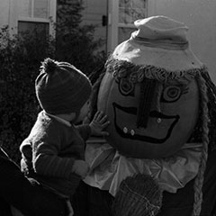 A mother holds her child in her arms in front of a scarecrow that has a decorated pumpkin with a grimacing face for a head.