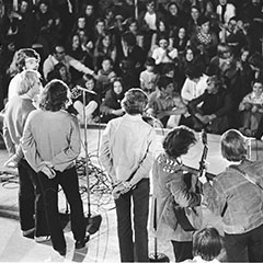 Seven musicians and singers perform on a stage in front of a crowd sitting on the ground.