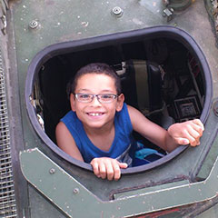 A smiling child coming out of a military tank's hatch.