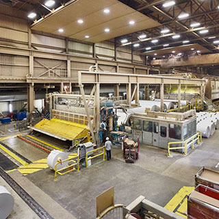 Interior view of a paper mill. Several large paper rolls are installed in the machine while others are lying on the floor.