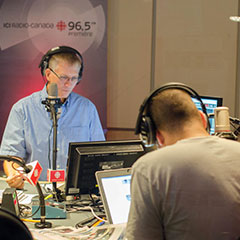 Studio de radio moderne où trois animateurs parlent aux micros avec de l'équipement informatique autour d'eux.