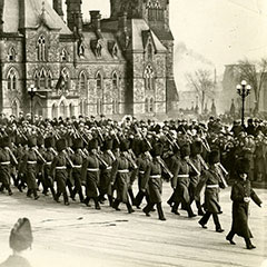 Soldiers from the Royal 22nd Regiment march in front of the Parliament building in Ottawa. A crowd watches them. In the foreground, we can see a man running.