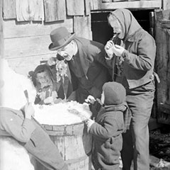 A man and woman with three children tasting maple taffy on snow in front of a wooden house.